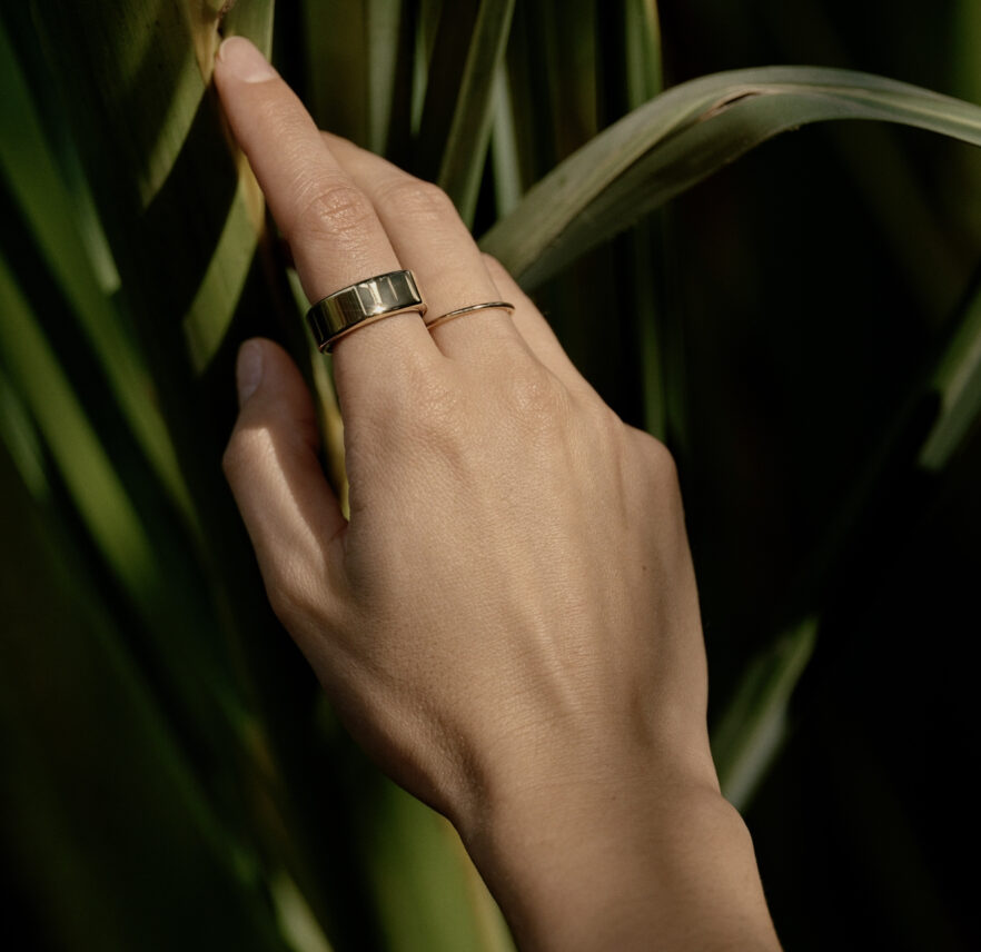 A feminine hand wearing an Oura Ring on the pointer finger, reaching into tall grass