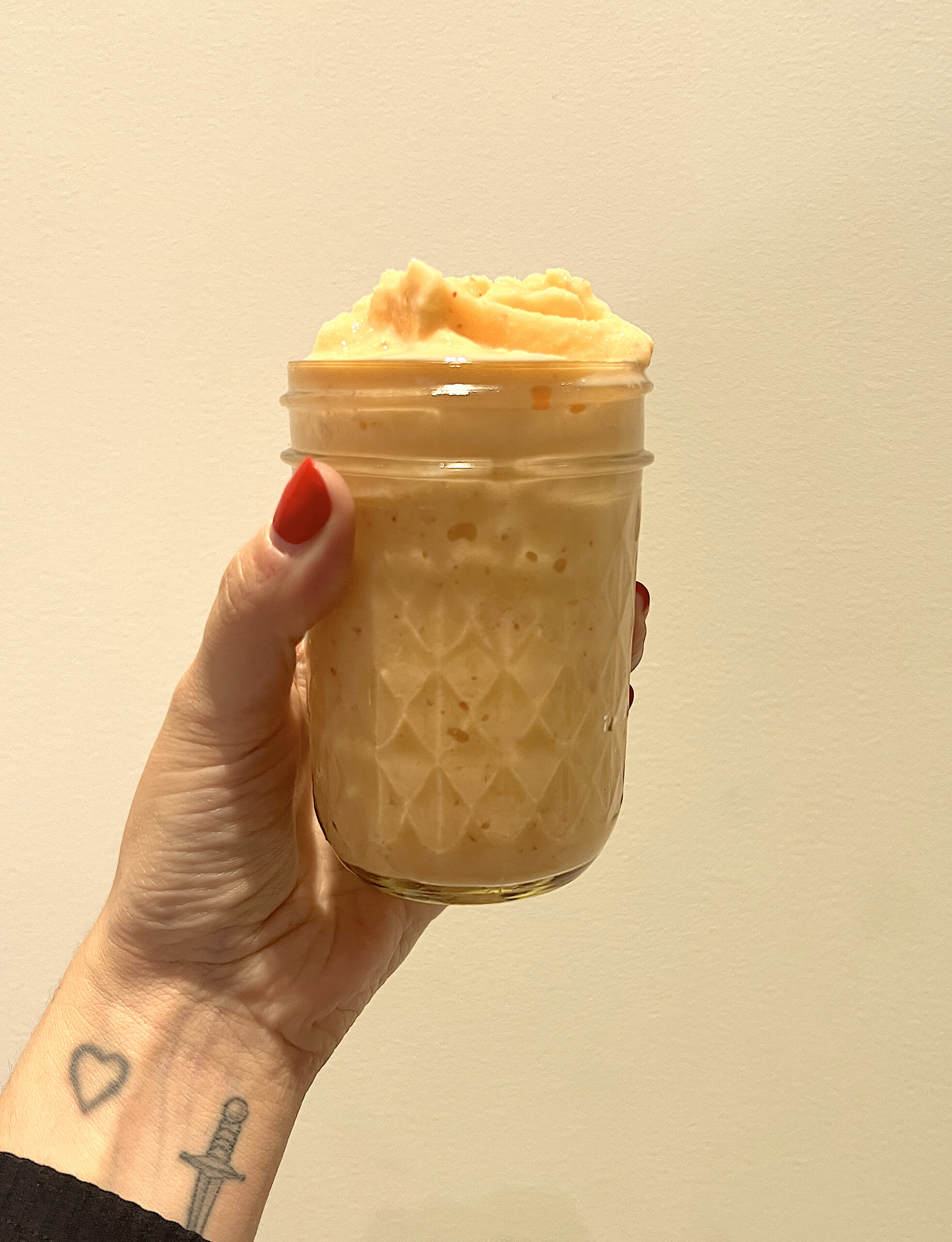A tattooed wrist and painted nails holds up a small orange smoothie in a glass jar