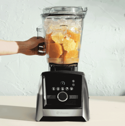 A Vitamix blender full of orange slices sits on a counter in front of a light blue background