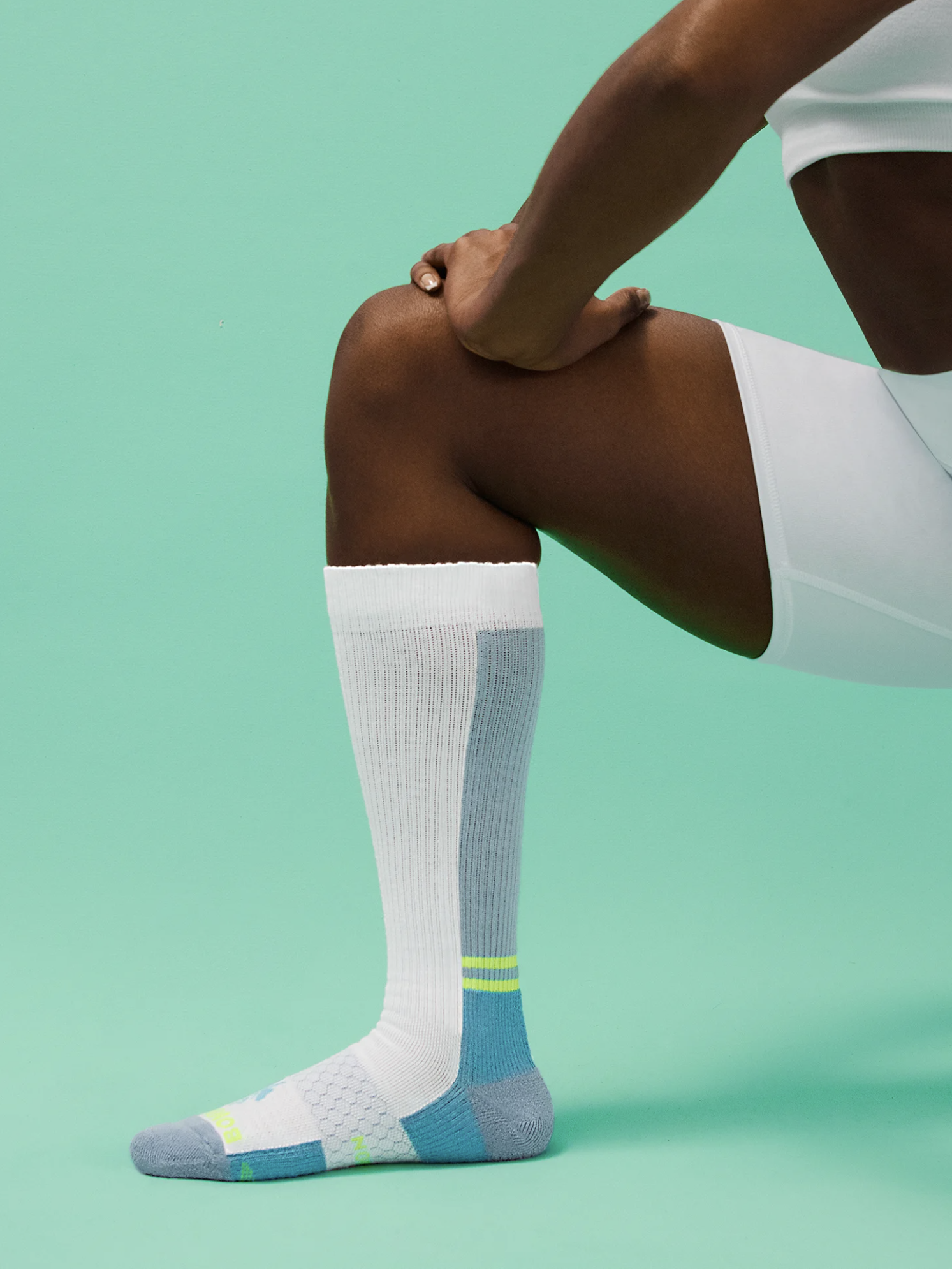 A person wearing white sportswear and a knee-high compression sock with a colored heel and toe, stretching the calf muscle against a green background.