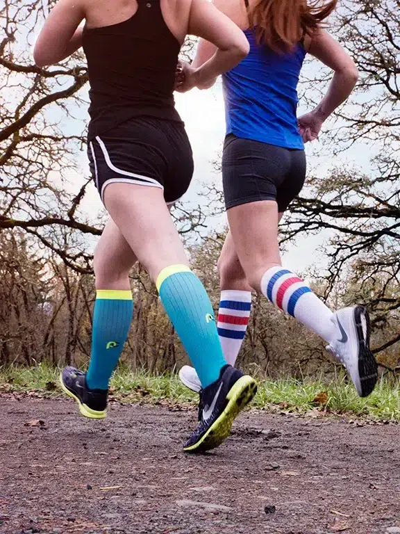 Two runners wearing compression socks and jogging on a dirt path.