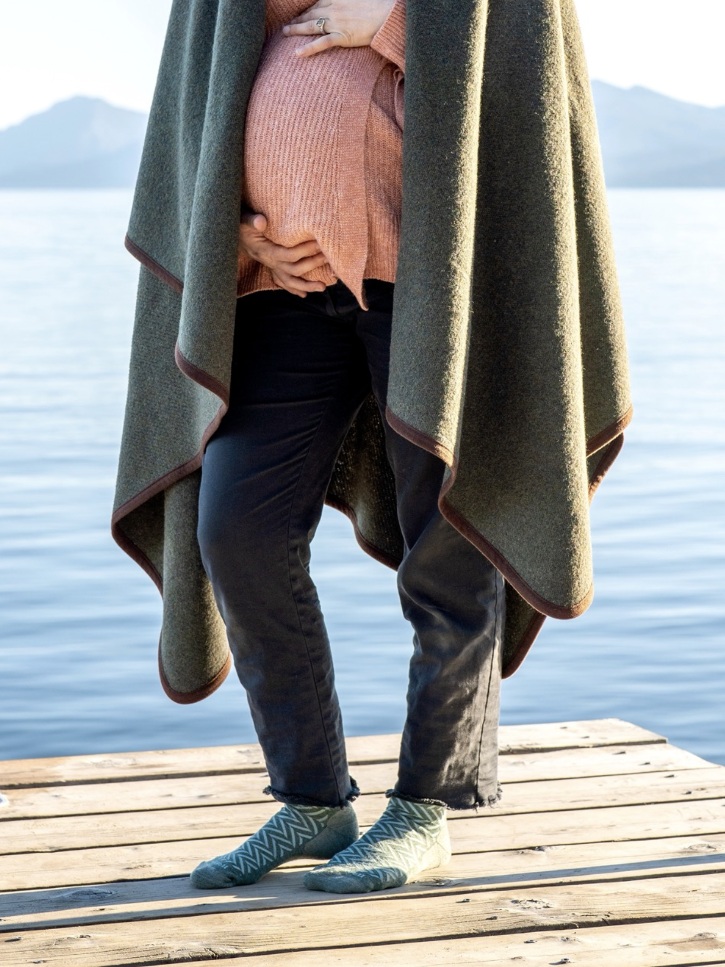 A person standing on a wooden dock by the water, wrapped in a green blanket and wearing black pants and patterned compression socks.
