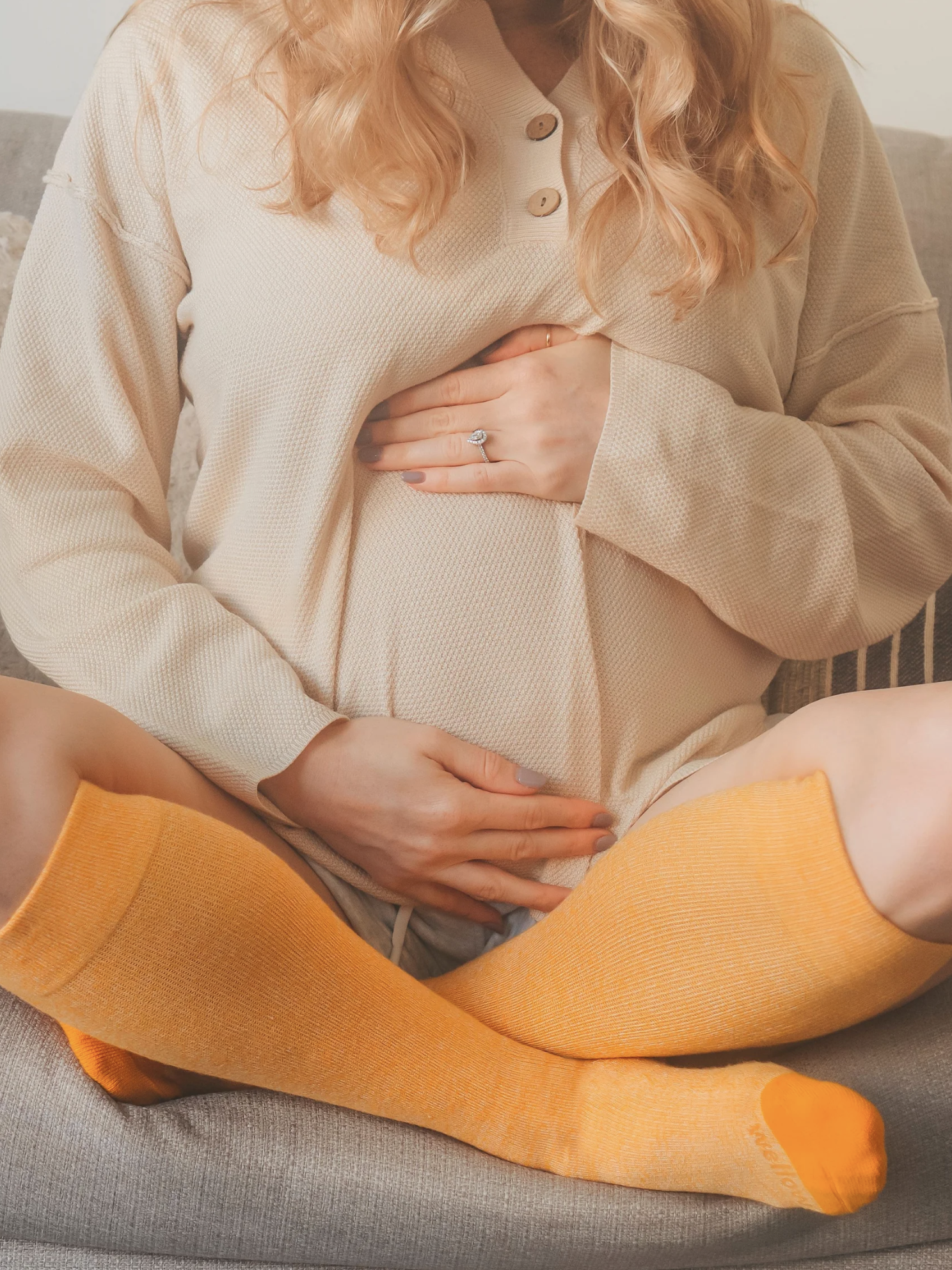 A person sitting on a couch wearing a beige top and orange compression socks, holding their pregnant belly.