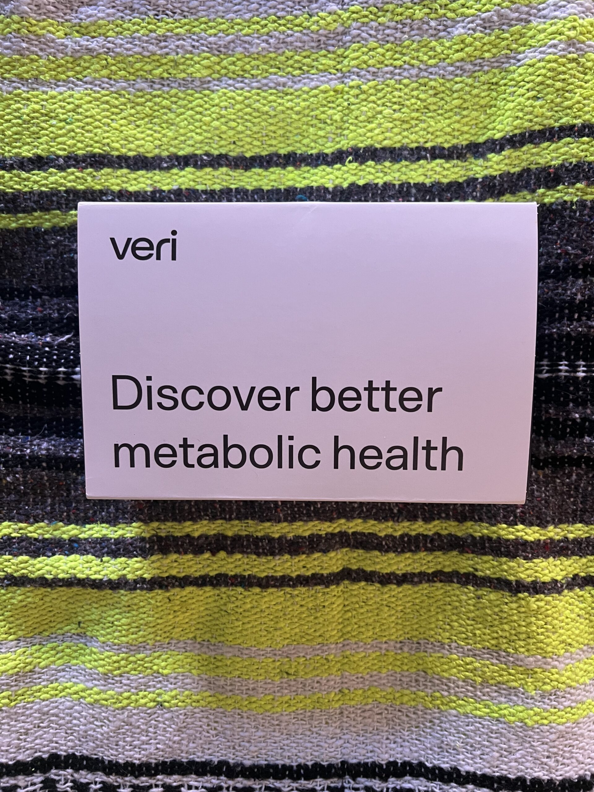 A sign with the text "veri discover better metabolic health" placed against a striped fabric background in shades of green and black.