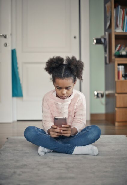 A young girl sits on the floor in the middle of her bedroom, looking down at a smartphone.