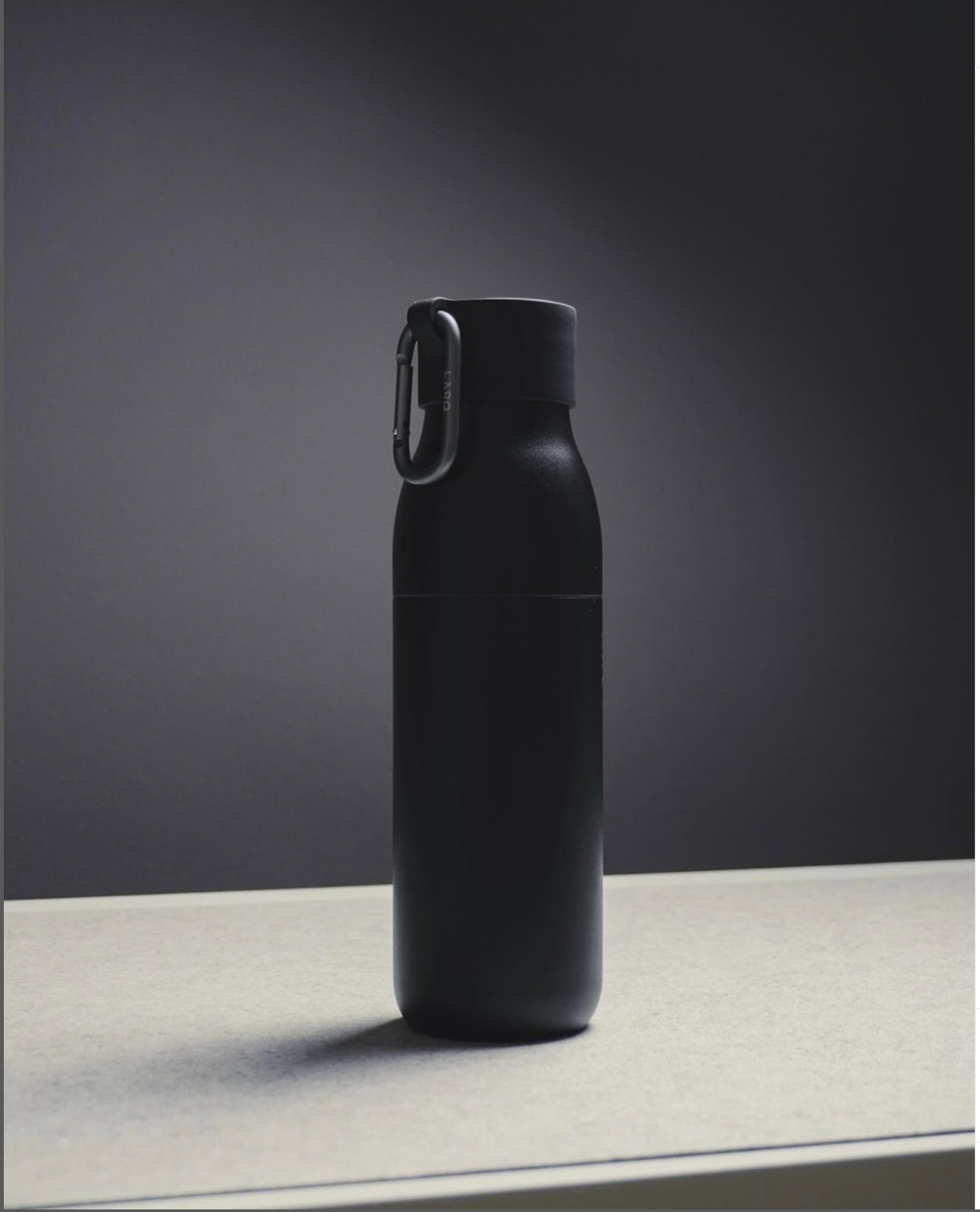 Black insulated water bottle with a carabiner attached to its lid, positioned on a table against a gray background.