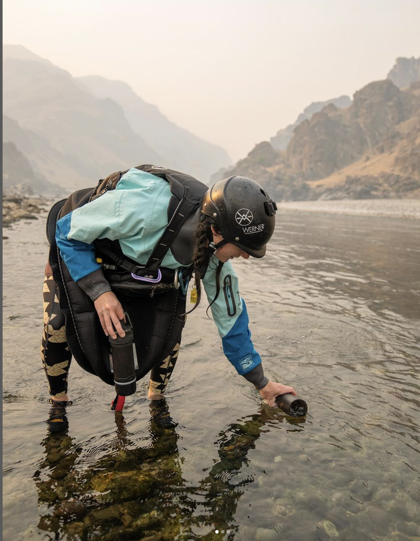 Researcher in protective gear collecting water samples from a river amidst hazy conditions, with an equipment bag.