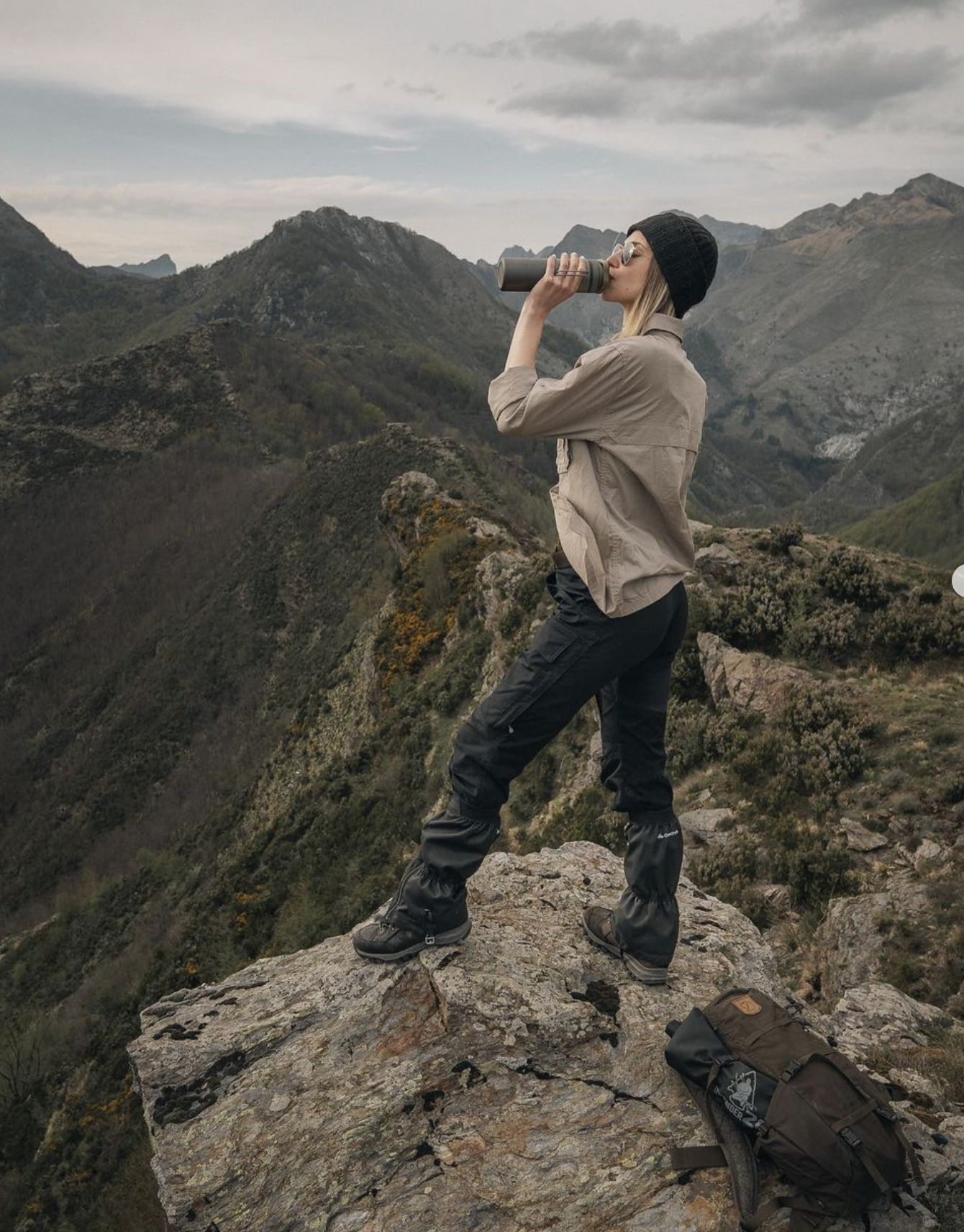 Person drinking from a bottle while standing on a mountain overlook with scenic hills in the background.