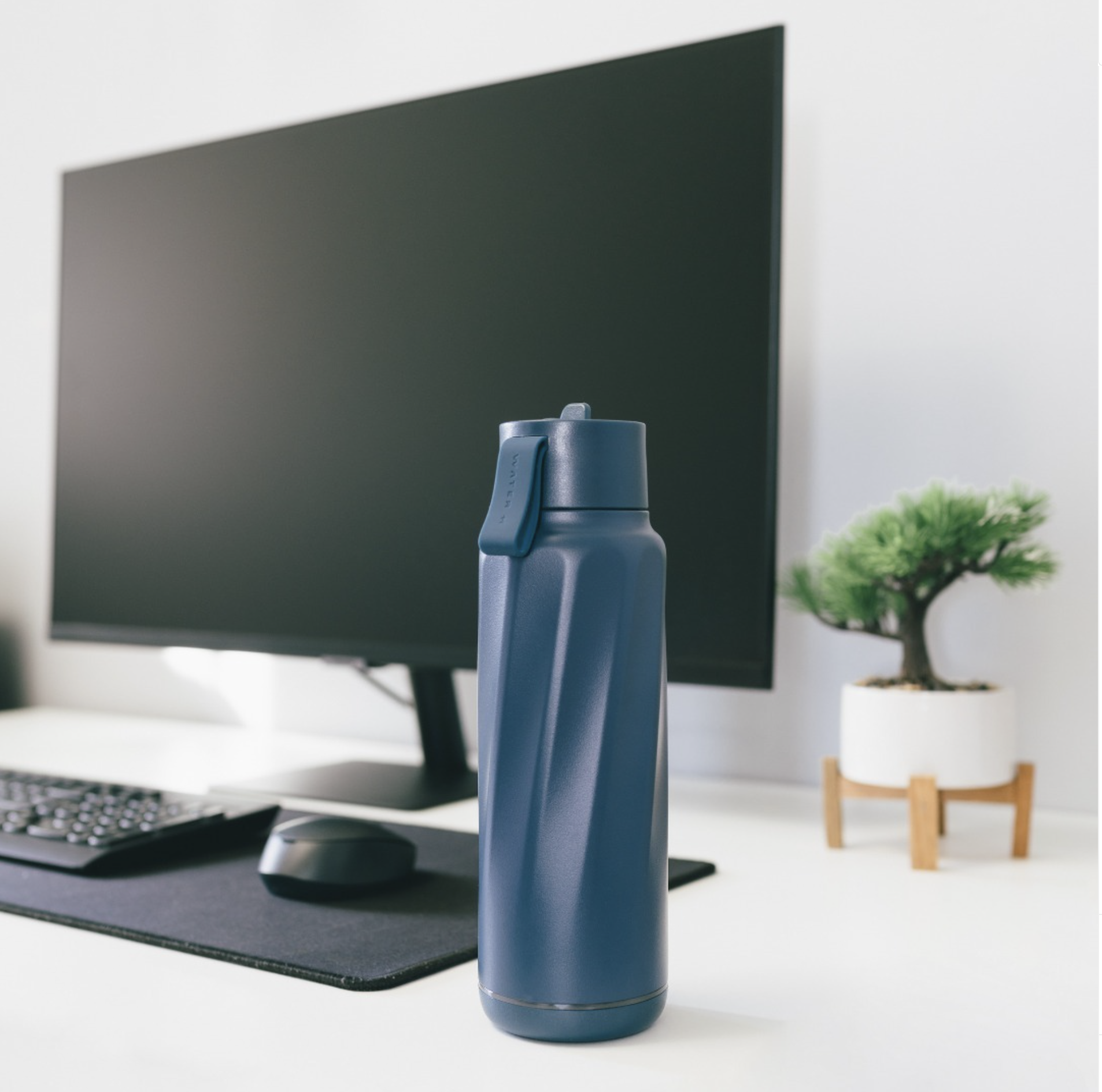 A blue insulated bottle placed on a desk in front of a computer monitor with a keyboard, and a mouse accompanied by a small potted plant.