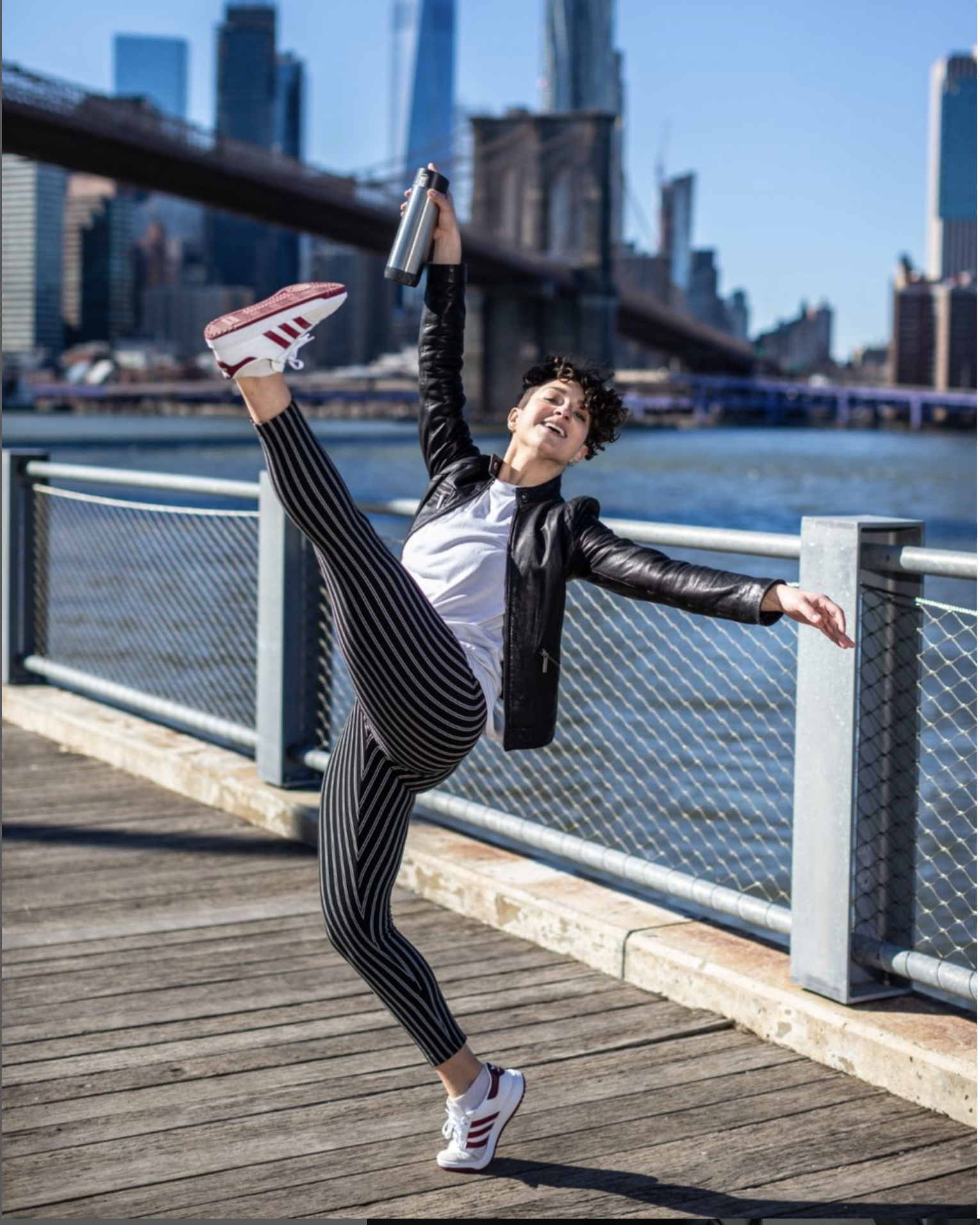 A person joyfully dancing on a sunny waterfront boardwalk with an urban skyline in the background.