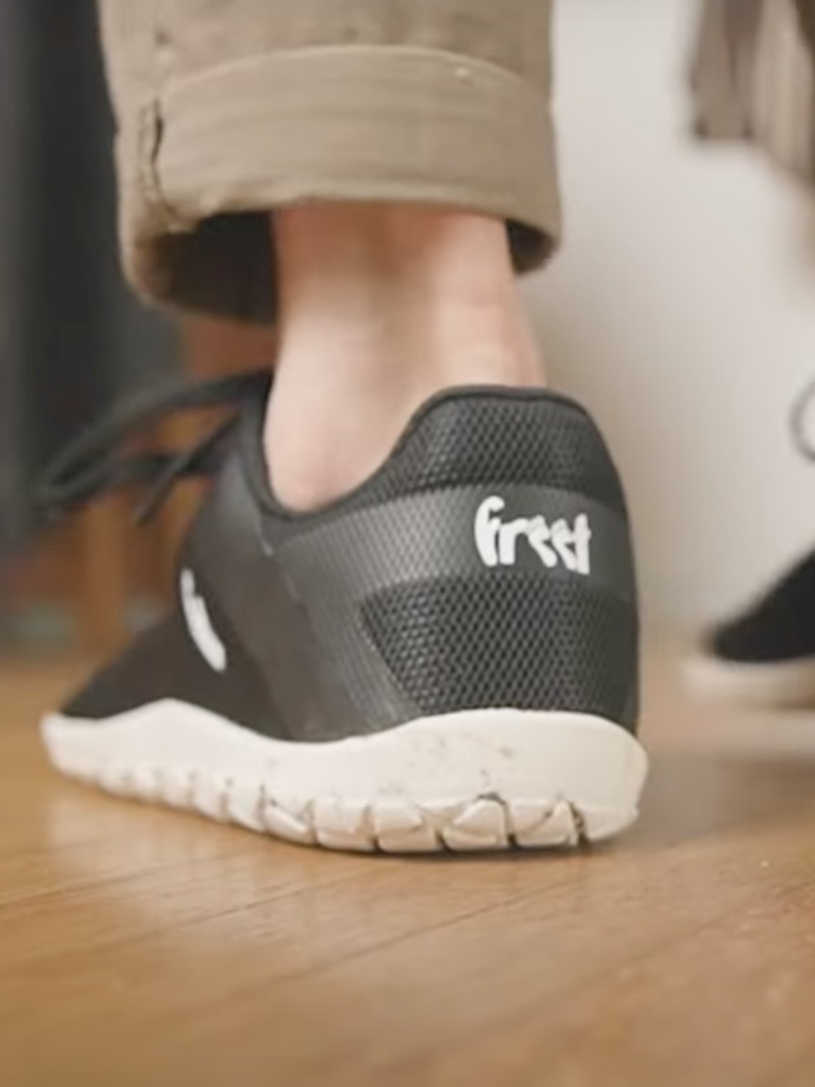 Person wearing black and white barefoot shoes with the word "first" written on the heel, stepping onto a wooden floor.