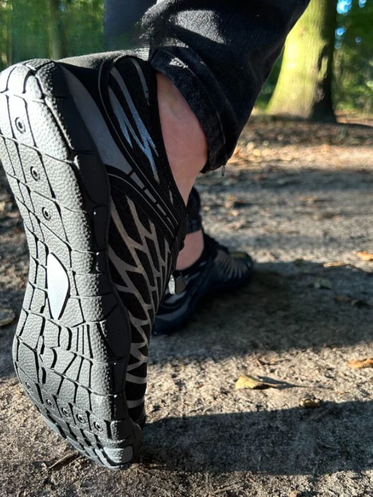 Close-up of a person's foot wearing a black barefoot shoe with a detailed tread pattern, stepping on a forest path.