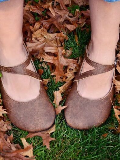 A pair of brown mary jane shoes on a woman's feet, standing on grass covered with fallen brown leaves next to massage guns.