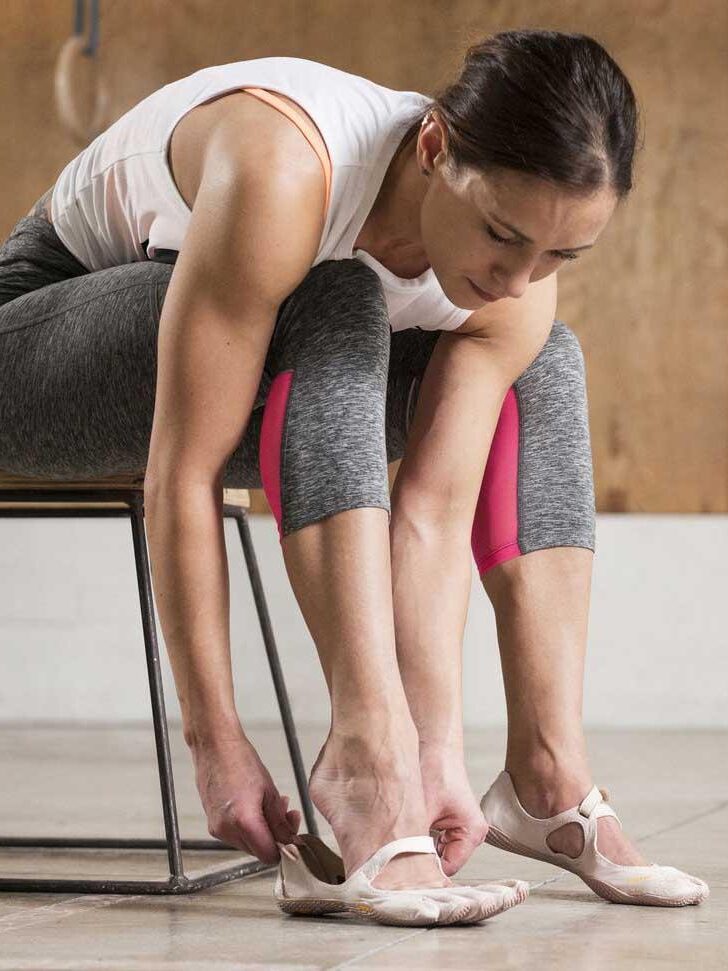 A female gymnast adjusts her foot straps on her slippers while sitting on a bench in a gym, beside her massage guns.