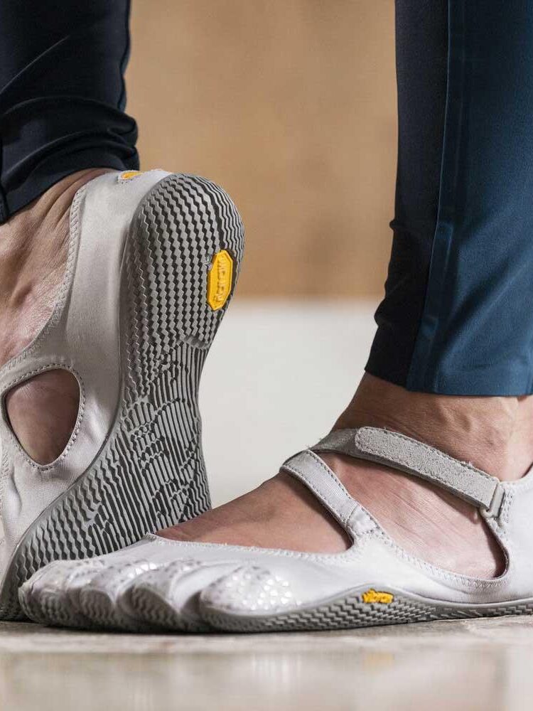Close-up view of a person wearing Vibram FiveFingers shoes in grey and white, standing on a beige tile floor, holding massage guns.