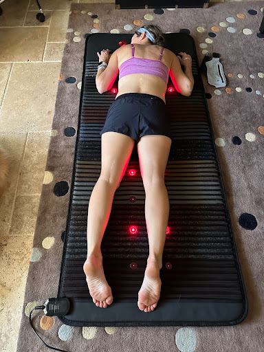 A woman lying on a red light therapy mat while wearing athletic shorts and a sports bra, with a smartphone beside her.