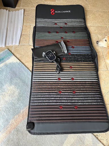 A black and red therapy mat with a controller placed on top of it, resting on a tiled floor next to a dog.