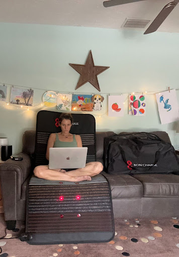A person sitting cross-legged on a massage chair using a laptop, with a room decorated with wall art and string lights in the background.
