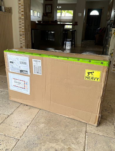 A large heavy package with shipping labels has been delivered and is sitting on a tiled floor inside a house.