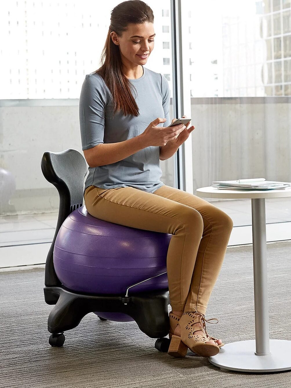 A woman sitting on a purple exercise ball chair, using a smartphone, in a modern office with large windows.