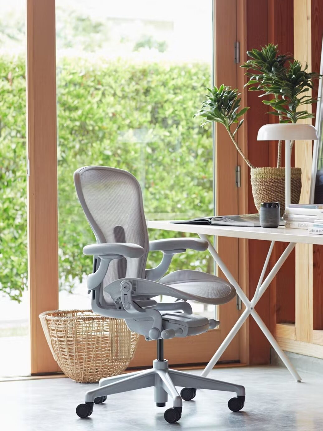 An ergonomic office chair next to a simple desk with a laptop, books, a potted plant, and a wicker basket, positioned by large windows and a view of greenery.