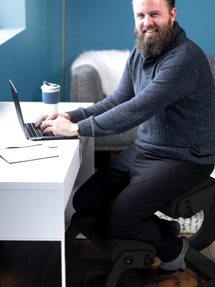 A smiling man with a beard working on a laptop at a white desk, wearing a gray sweater, black pants, and seated on a modern chair.