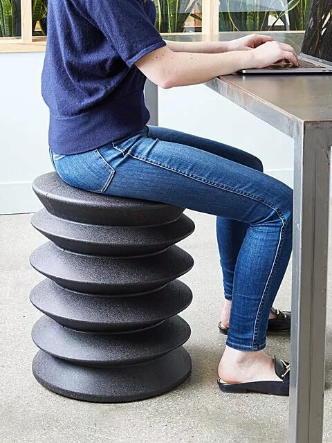A person in jeans and a t-shirt sits on a unique, spiral-shaped black stool while typing on a laptop at a metal table.