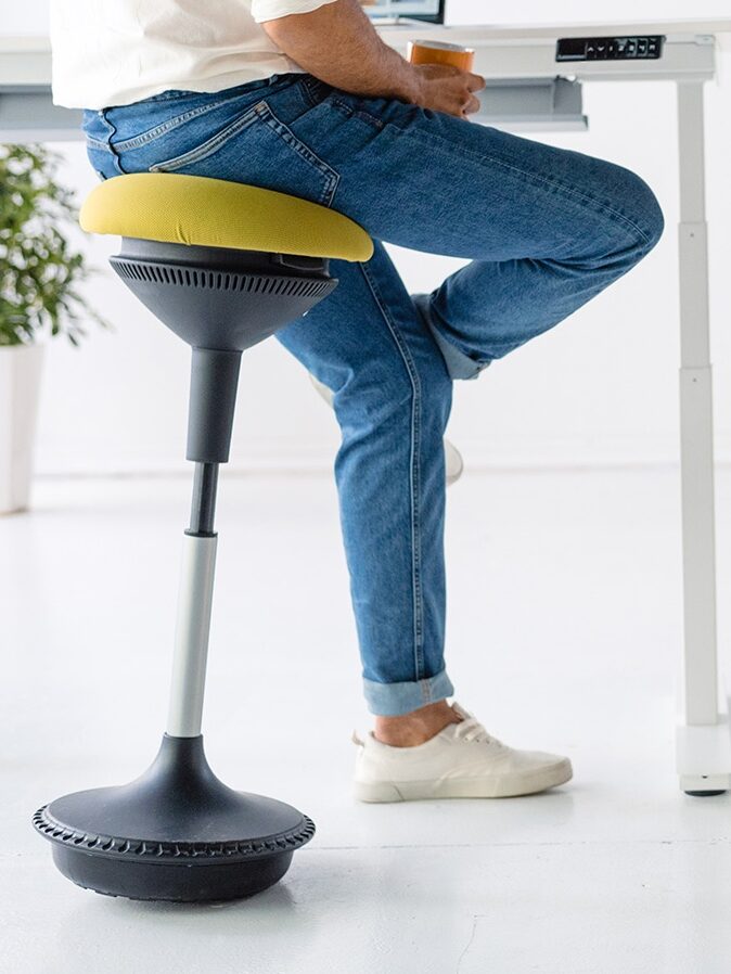 A person in jeans and sneakers sitting on a modern ergonomic stool in an office with a white desk and a clean, minimalist background.