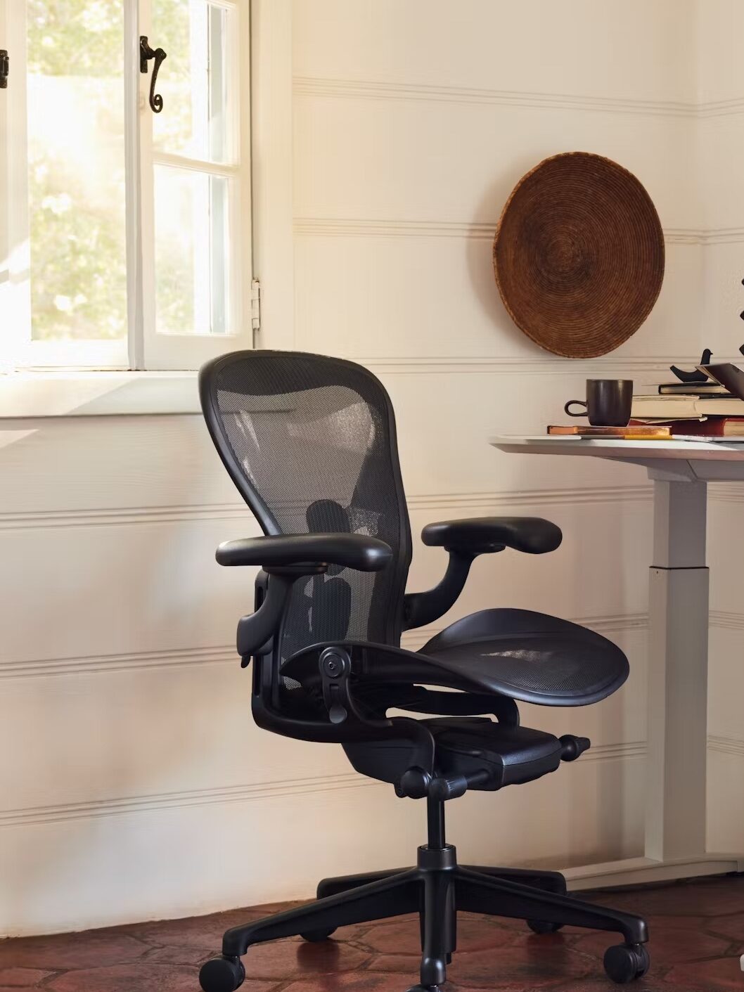 An ergonomic office chair with adjustable armrests in a bright room with a wooden desk and terracotta floor.