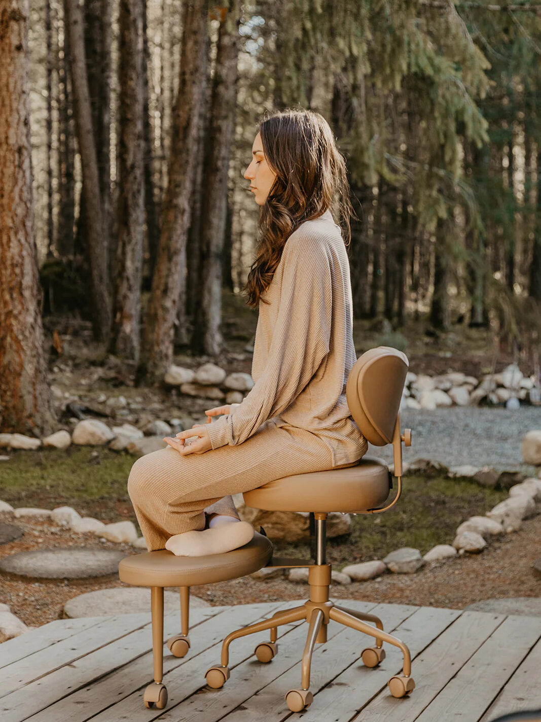 A woman in a beige outfit sits on a rolling chair on a wooden deck, surrounded by a forest and sunlight filtering through the trees.