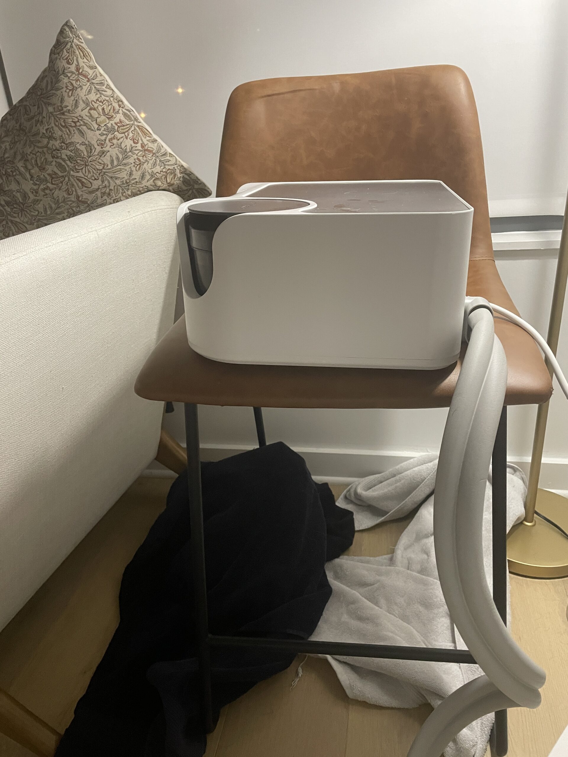 A white electronic device with hoses is placed on a brown chair. Nearby is a section of a beige couch, black bag, white towels, and a floor lamp.