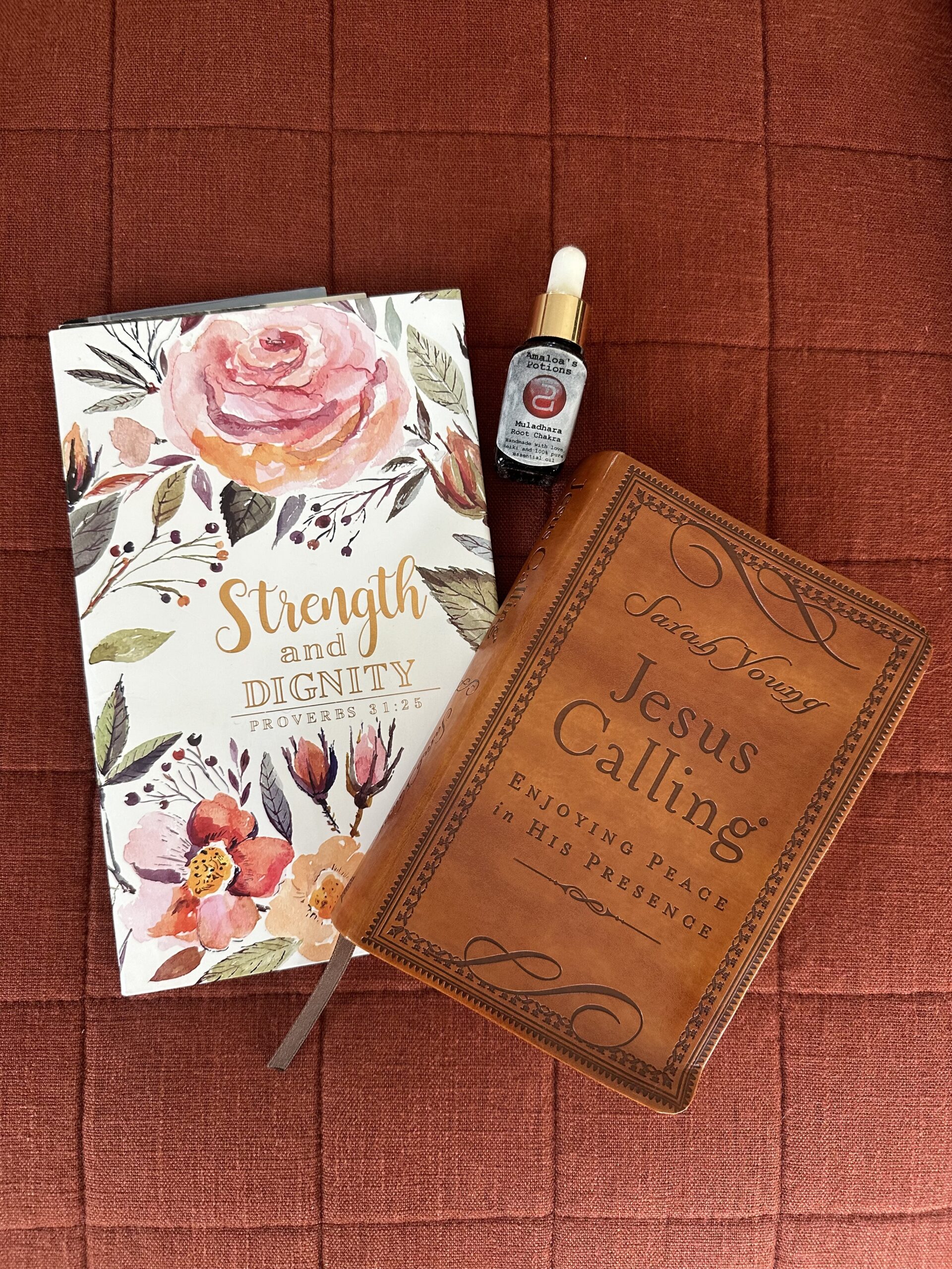 A floral journal, a book titled "Jesus Calling," and a small bottle of essential oil are placed on a textured orange surface.