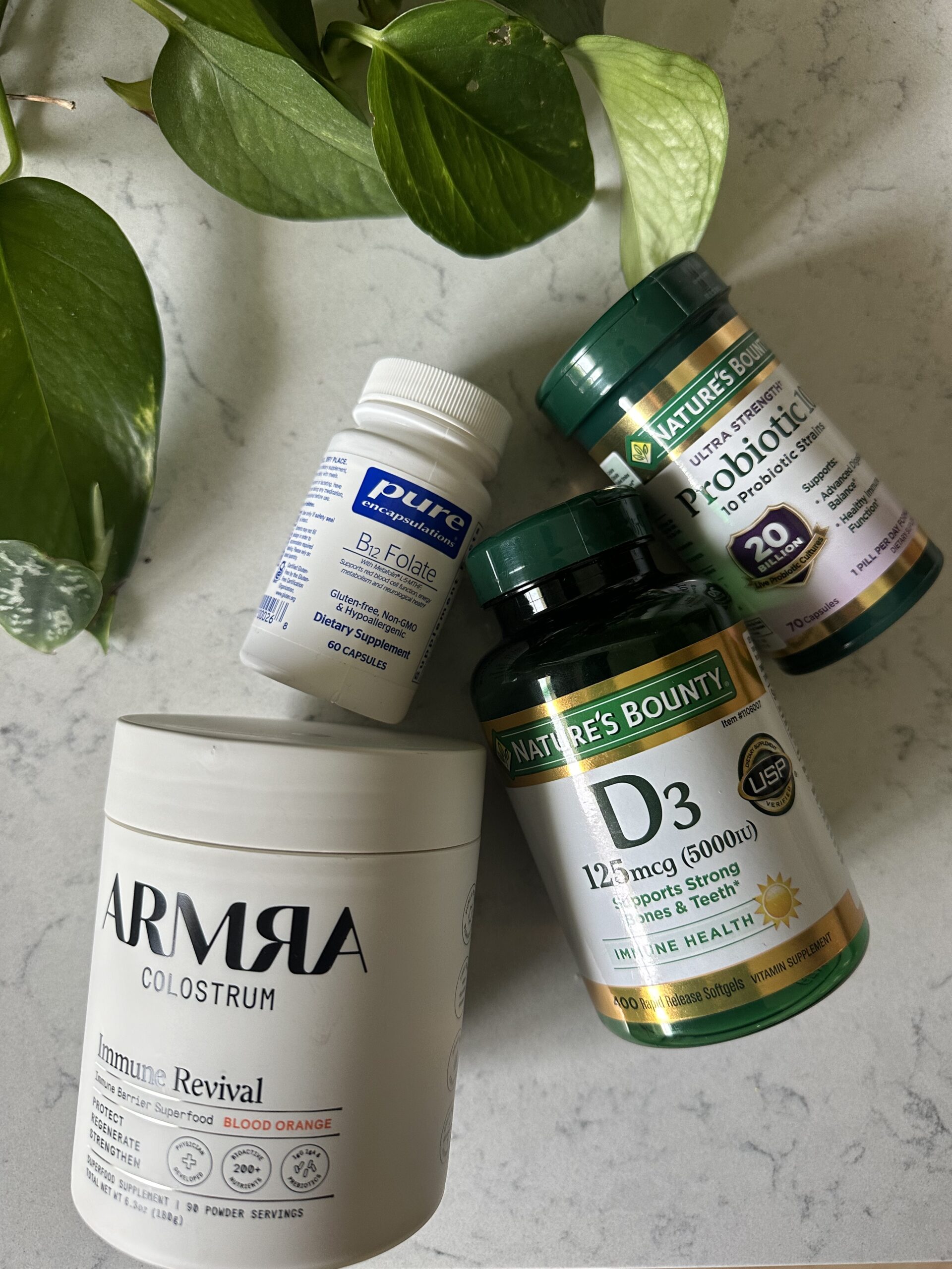 Bottles of supplements including B-Folate, probiotics, vitamin D3, and colostrum are placed on a marble surface with a leafy plant nearby.