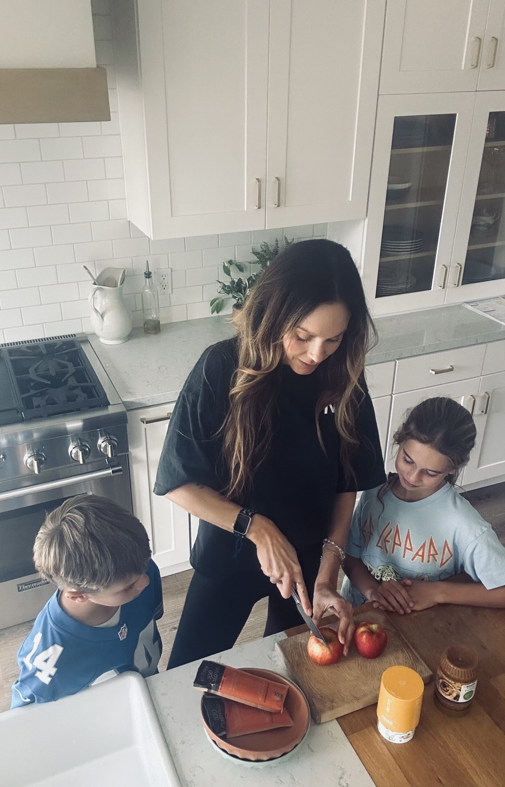 A woman slices apples on a cutting board in a kitchen, with two children watching nearby.