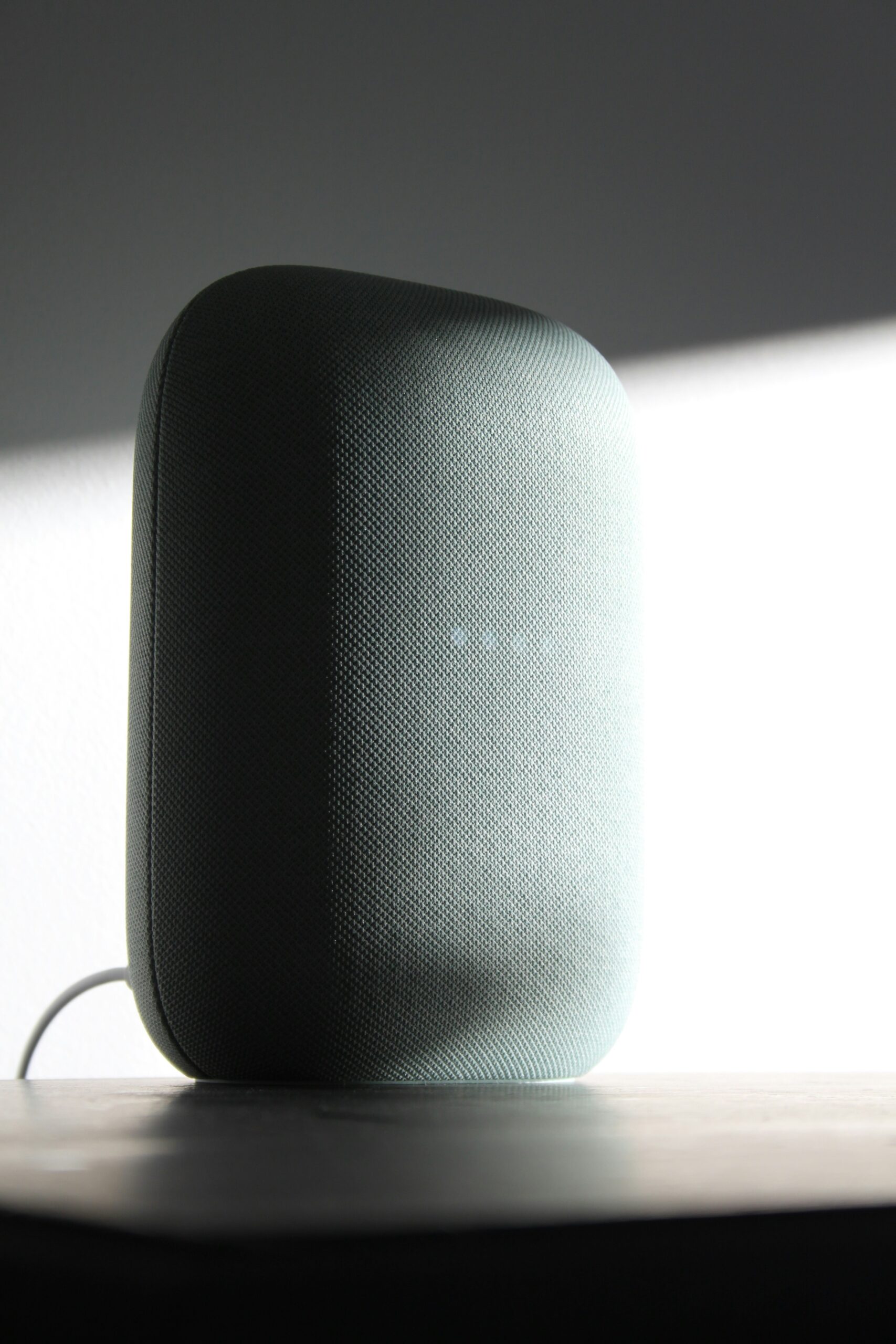 A smart speaker with a dark fabric cover sits on a wooden surface, backlit by bright light.