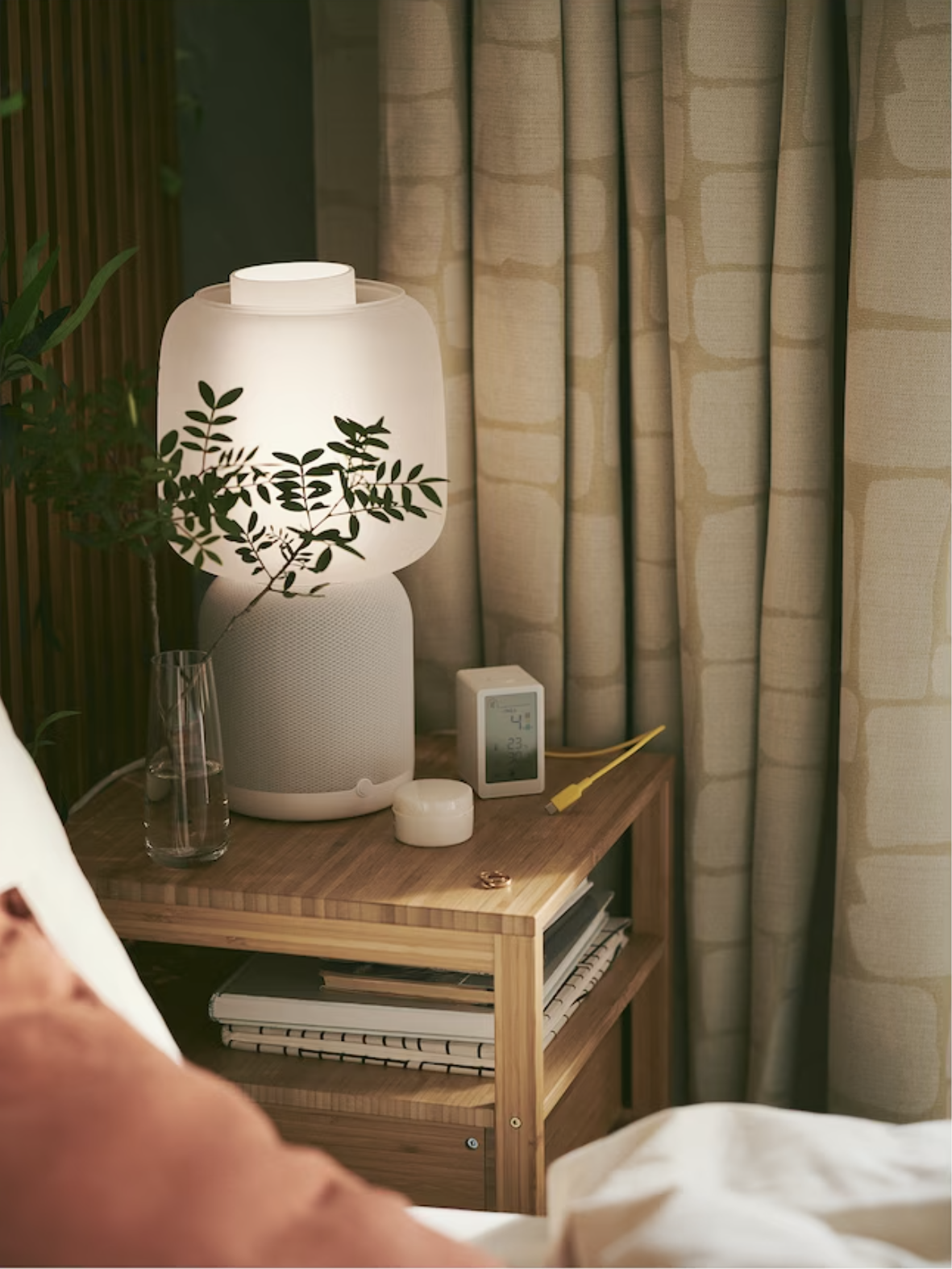 A bedside table with a modern lamp, small clock, and a vase with greenery. Nearby, a bed with a pillow offers comfort, while curtains hang in the background.
