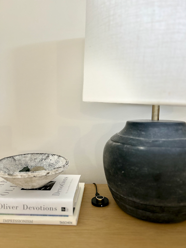 A ceramic lamp, a rough-textured bowl, and two stacked books sit on a wooden surface with a small black oura ring holder beside them.