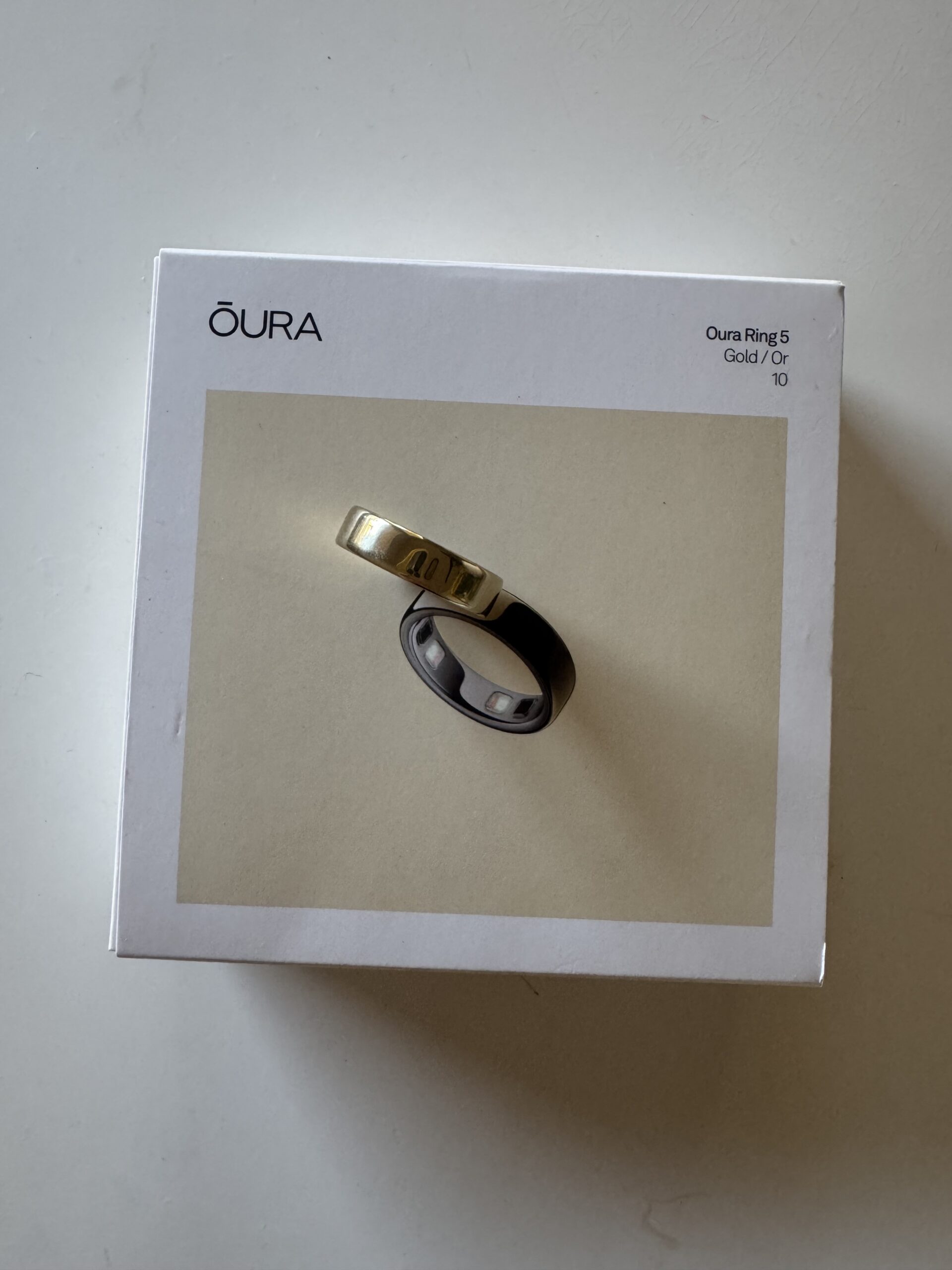 A box labeled “Oura Ring 3 Gold” featuring an image of two Oura rings—one gold, one black—prominently displayed on the cover, sits atop a white surface. This packaging highlights the elegant design of the Oura ring collection.