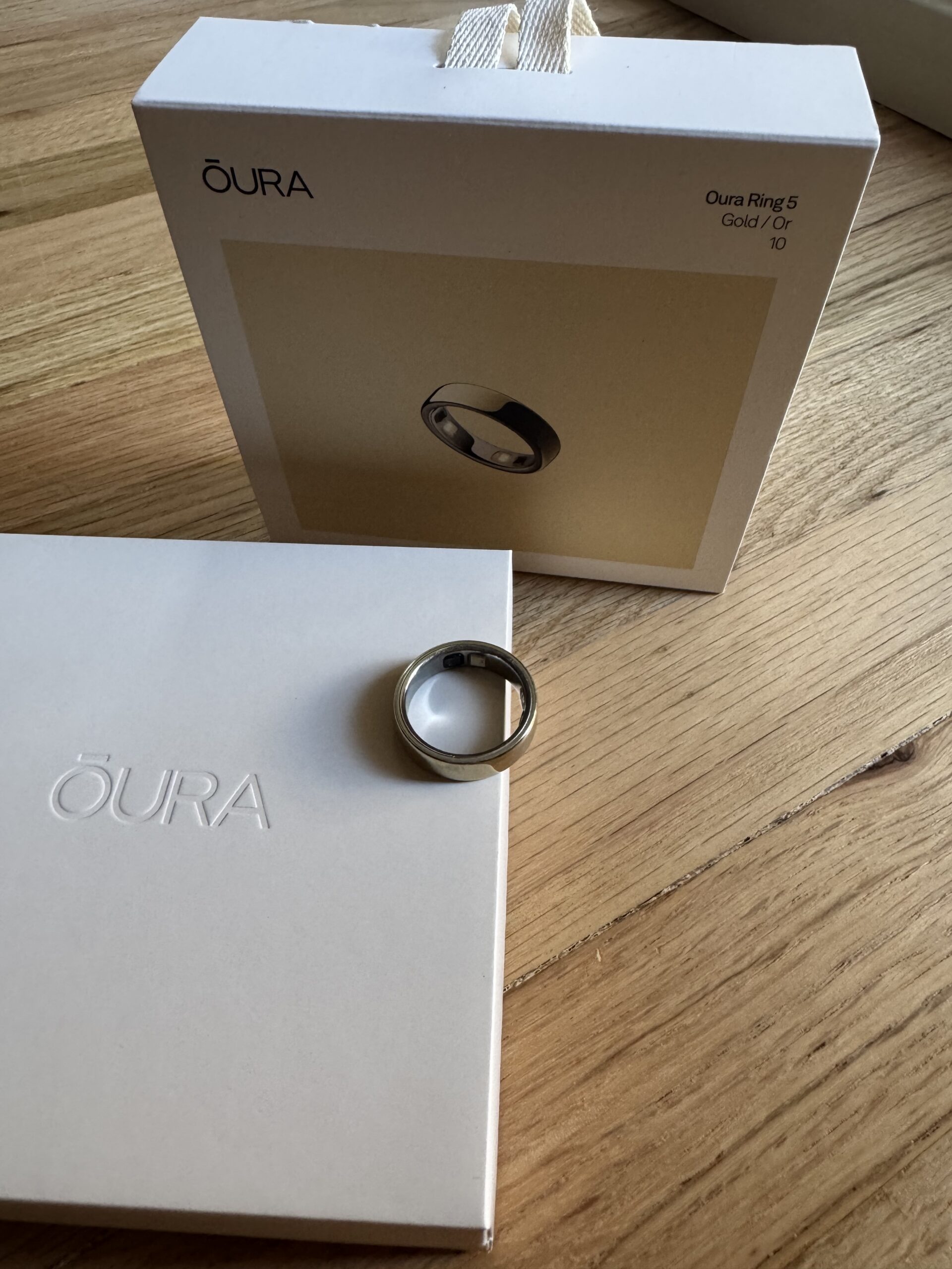 Oura Ring 5 in gold color, size 10, displayed on top of its packaging with the Oura Ring product box in the background, on a wooden surface.