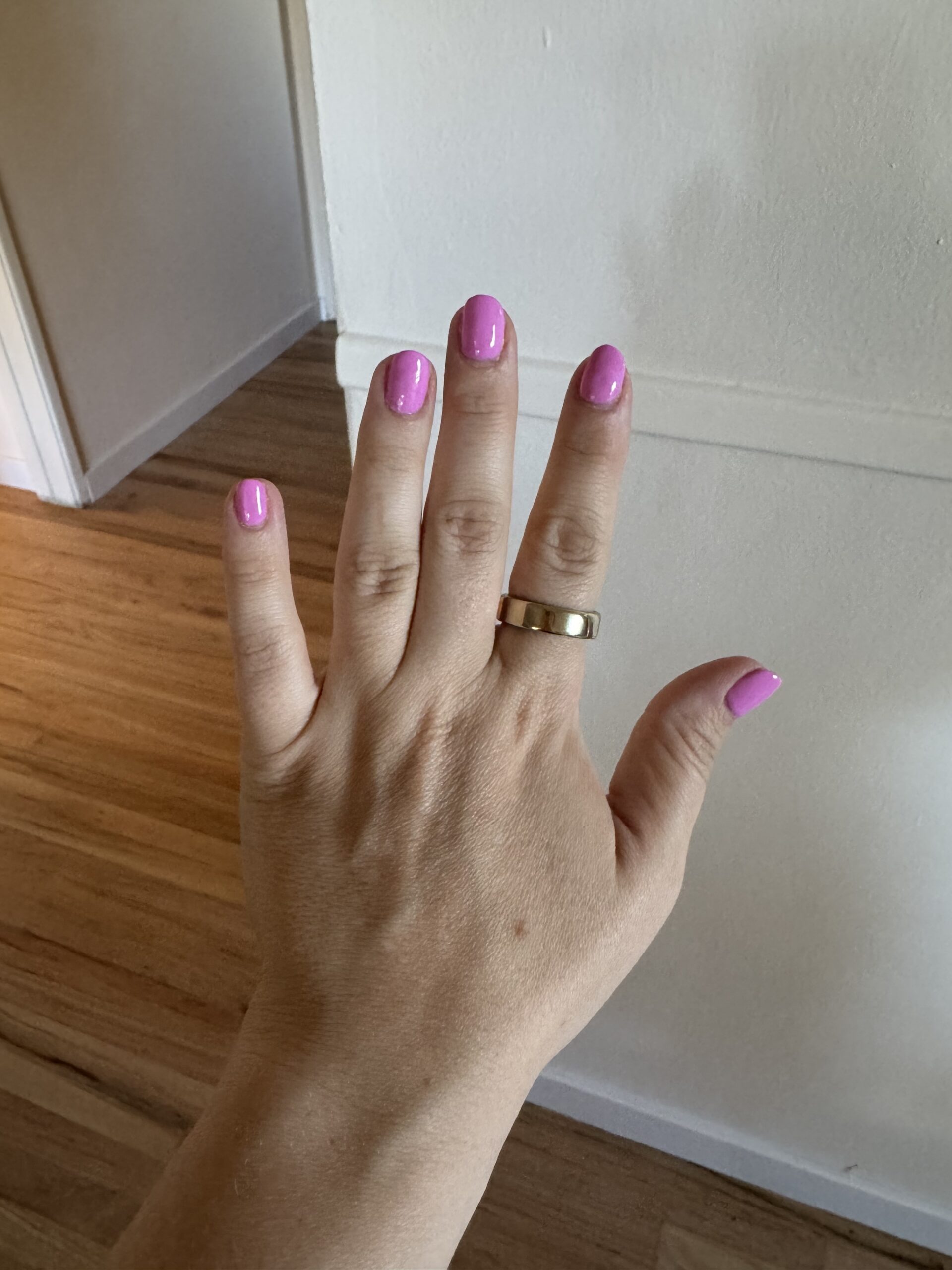 A hand with short, glossy pink nails and an Oura ring on the ring finger is held up against a pale wall and wooden floor background.