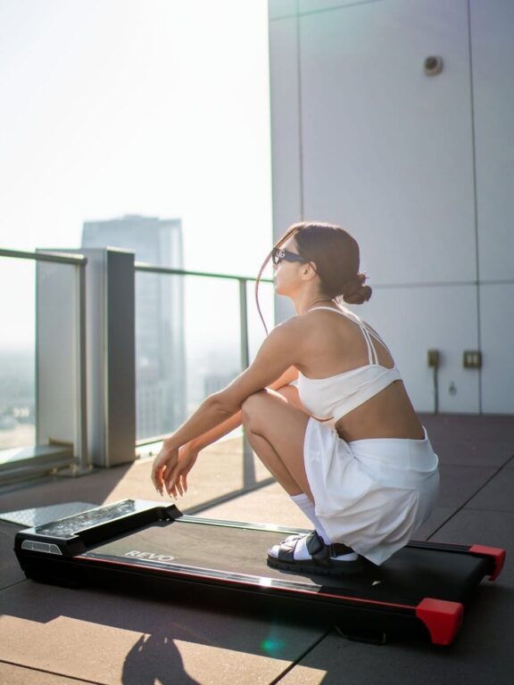 A woman in white workout clothes squats on a treadmill on a rooftop balcony with a city skyline in the background.