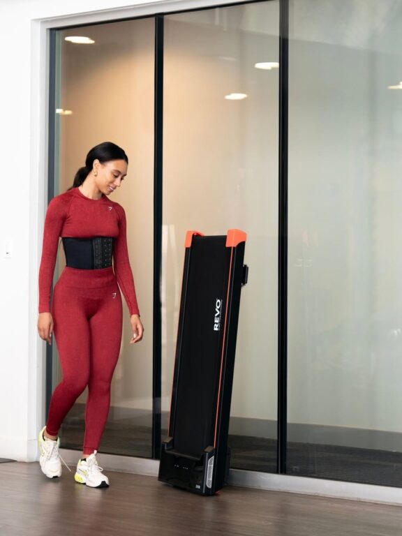 A woman in red workout attire stands indoors beside a folded black and orange RLVO treadmill leaning against a glass wall.