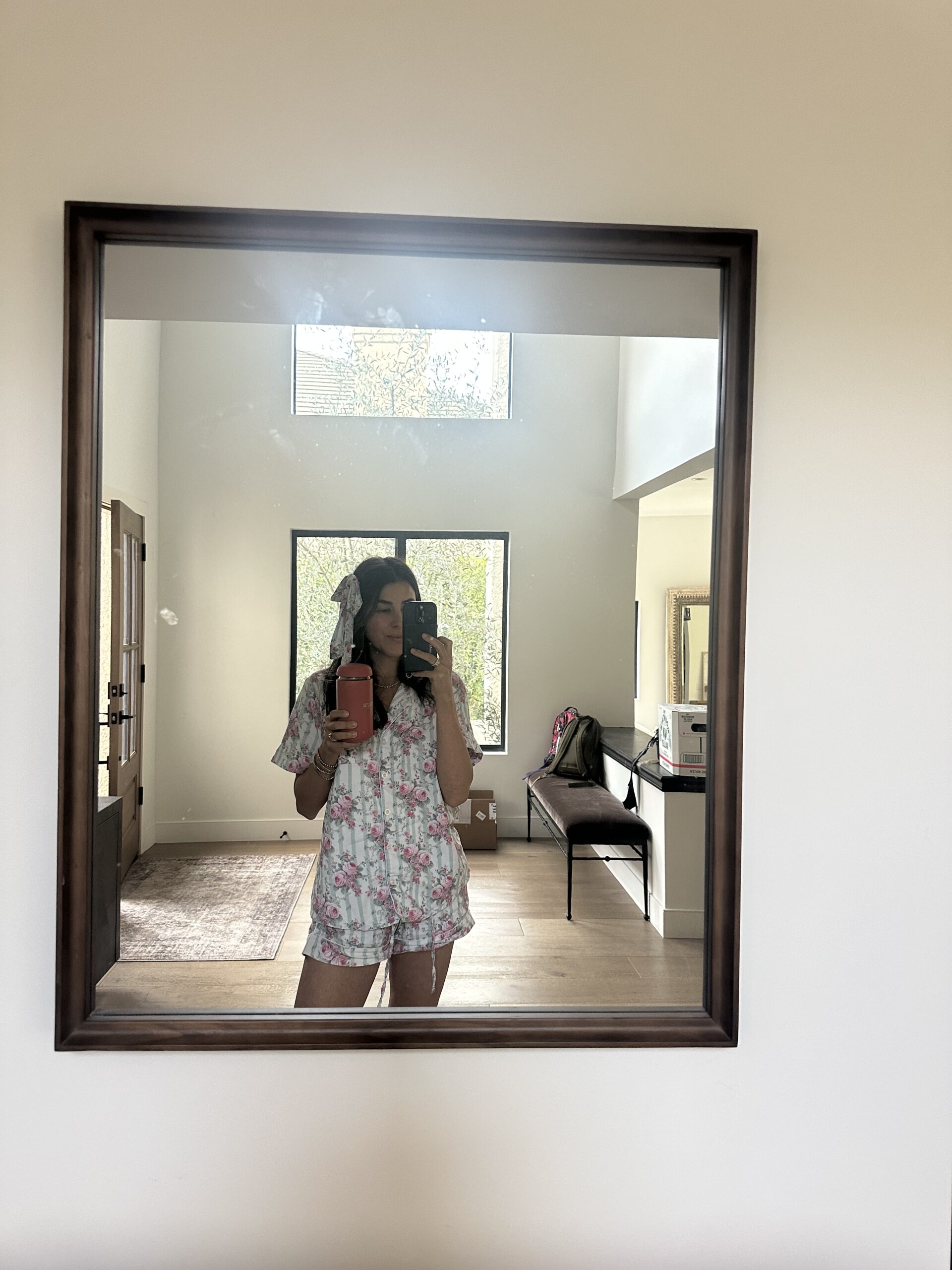 A person in floral pajamas takes a mirror selfie in a bright room holding a pink drink. A backpack and other items are visible in the background.