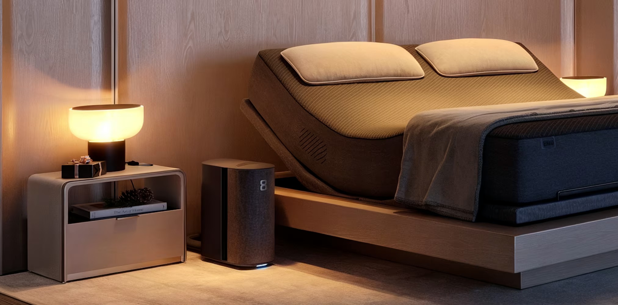 A modern bedroom with a beige adjustable bed, two pillows, a gray blanket, two bedside tables with lamps, and an Eight Sleep air purifier on the floor for a restful environment.