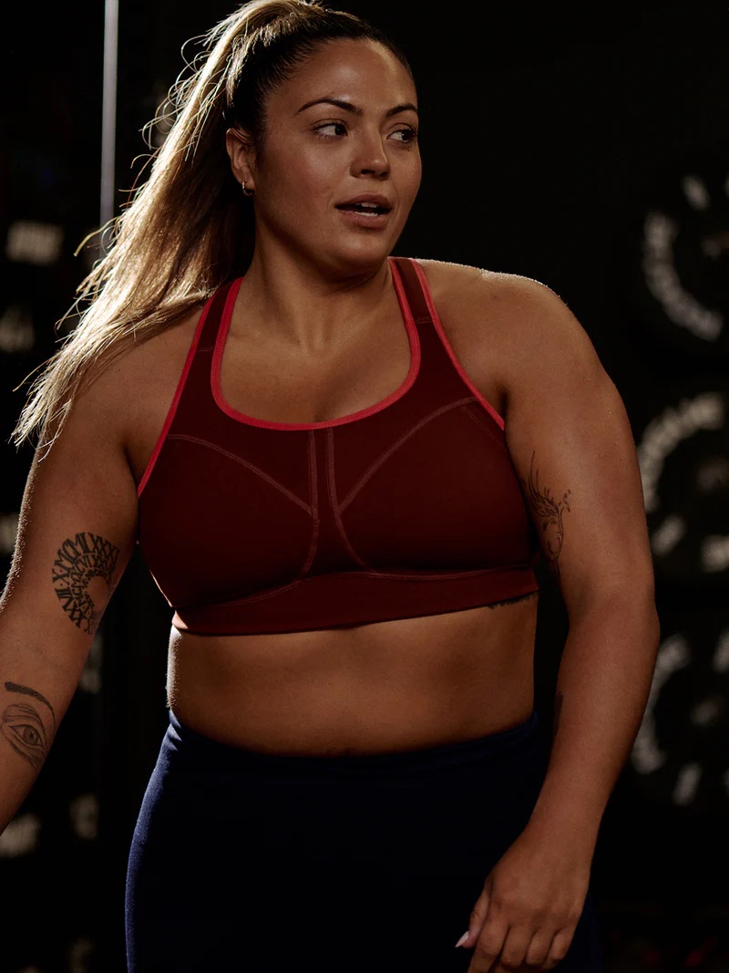 In a gym setting, a person in a dark red sports bra and pants gazes sideways with a neutral expression, tattoos adorning their arms. Hair tied back, they consider incorporating massage guns into their routine for optimal recovery.
