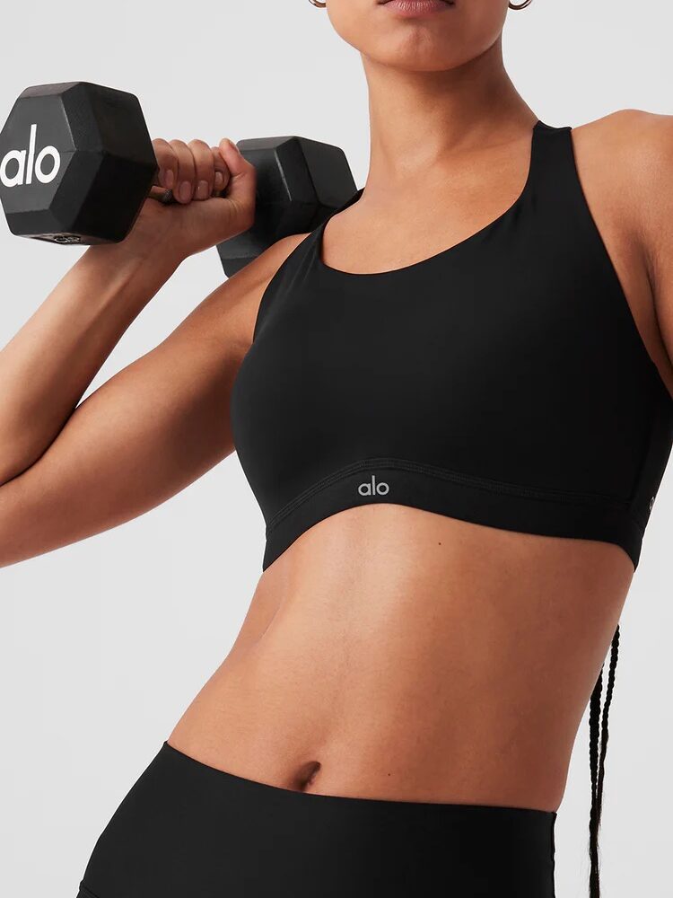 A person in a black sports bra holding a dumbbell over their shoulder, with "alo" visible on the bra and dumbbell, seems ready for an intense workout. Nearby, massage guns await to aid muscle recovery post-exercise.