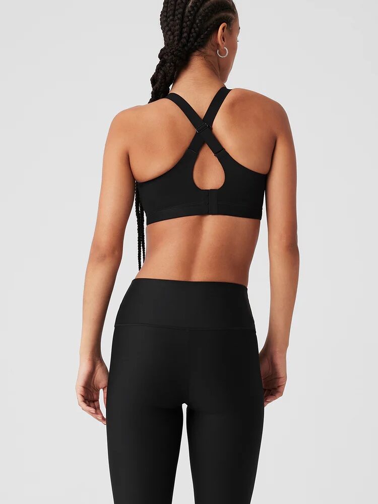 A person in black athletic wear stands against a plain background, showcasing a sports bra with crisscross straps and leggings. Nearby, massage guns are poised for post-workout recovery.
