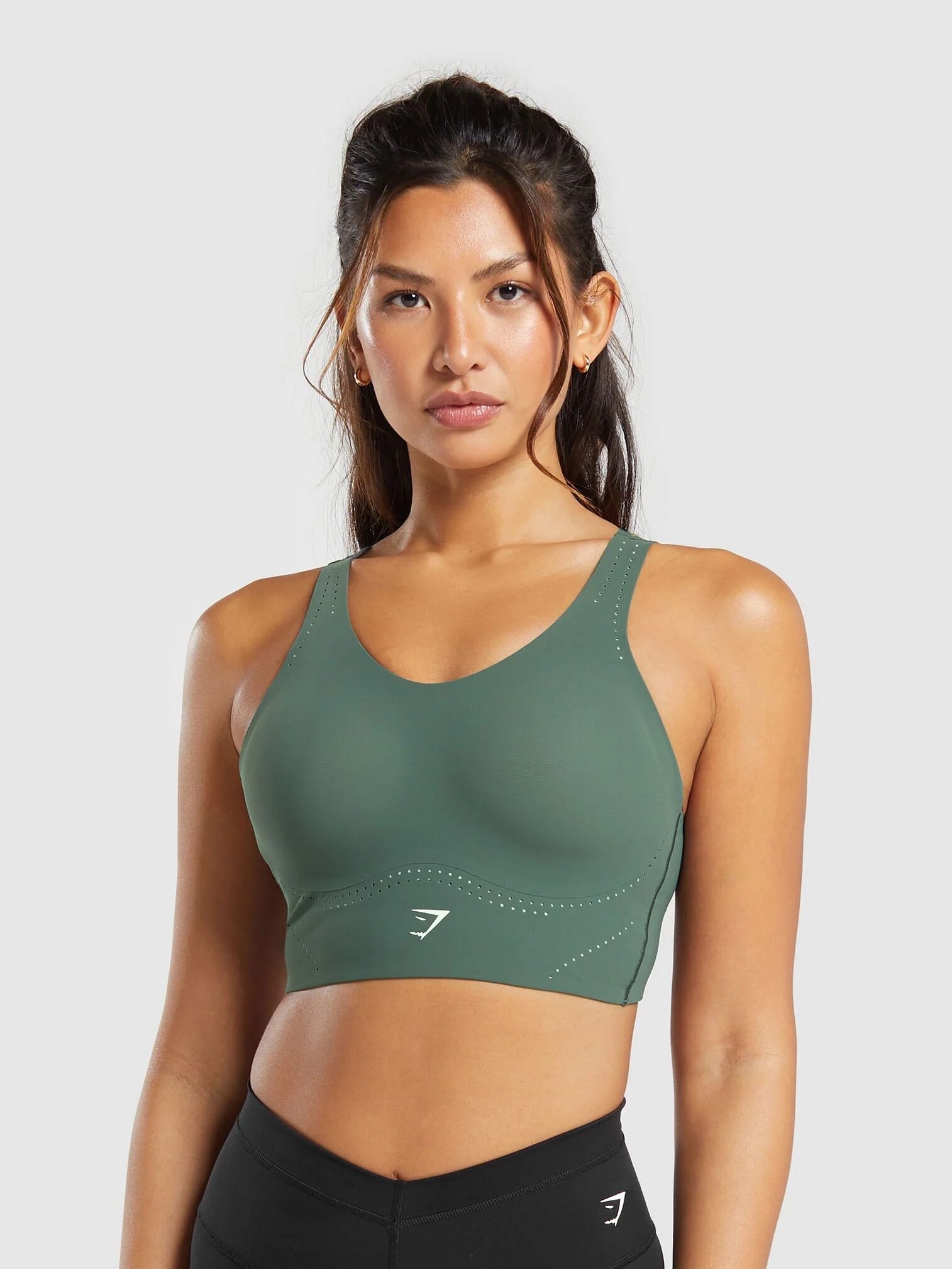 A person wearing a green sports bra and black leggings stands against a plain background, holding one of the latest massage guns.