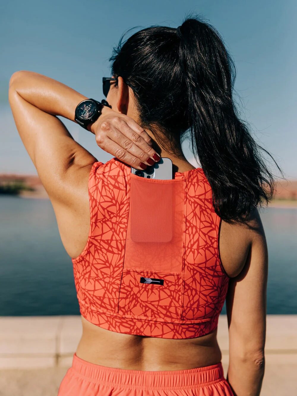 A woman in an orange sports outfit carries a phone in a pocket on her back, hand resting on her neck, possibly pondering the soothing relief of massage guns. She stands by a body of water on a clear day.