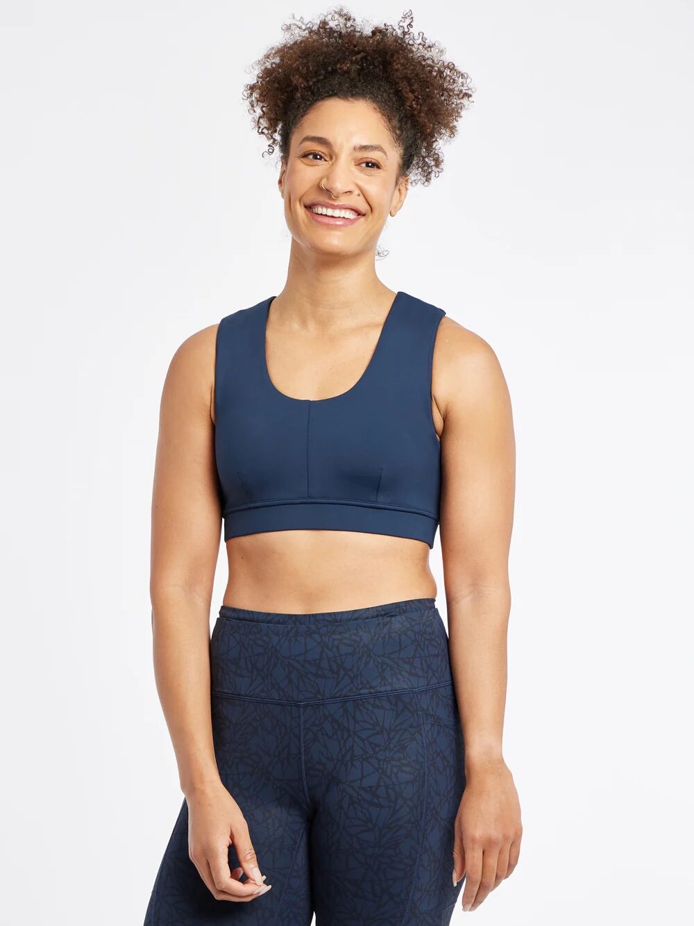 Person smiling, in a blue sports bra and patterned leggings, stands confidently with a massage gun in hand against a plain white background.